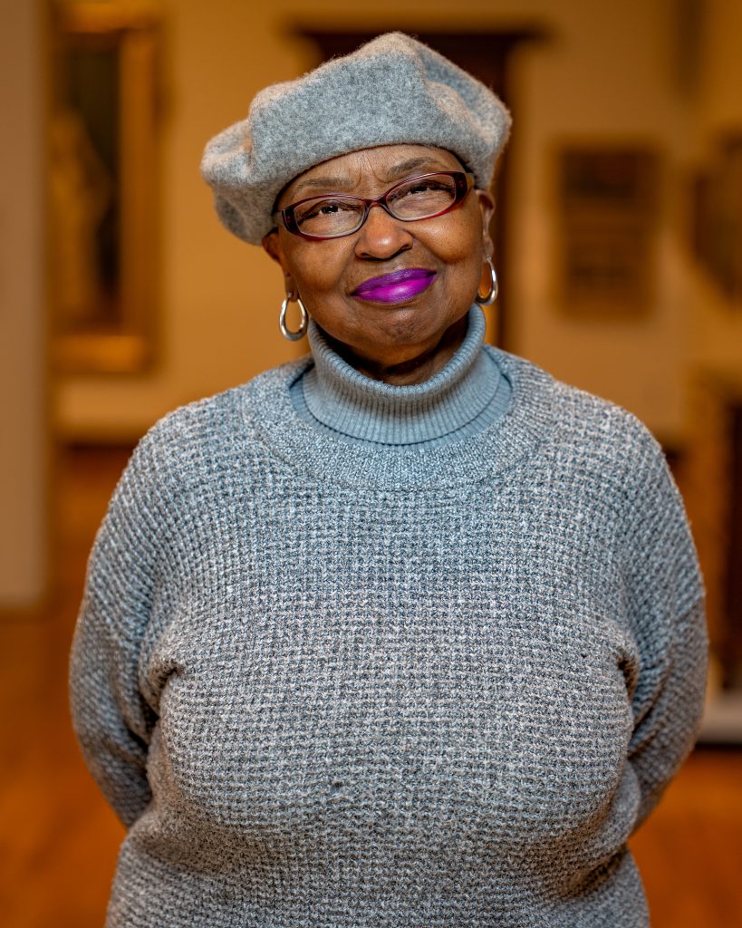 Barbara Hepburn in a grey shirt and hat with glasses and pink lipstick